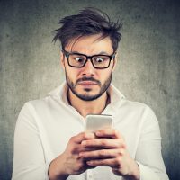 Young bearded man in glasses watching smarpthone and looking scared with bad news.