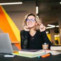portrait of young pretty woman sitting at table in black shirt working on laptop in co-working office, wearing glasses, having idea